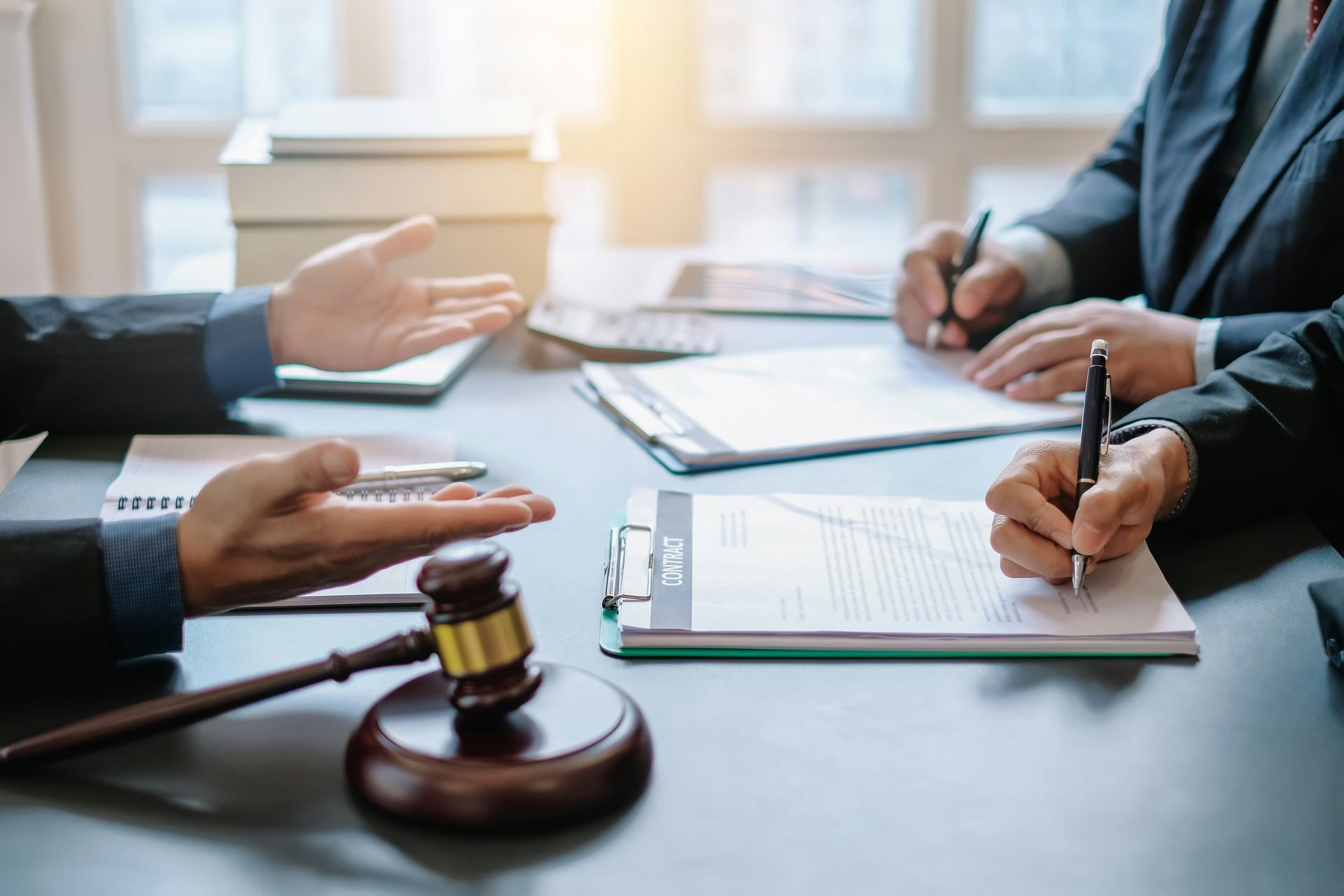 Three people in suits at a table with documents, gavel, and notepad, discussing and writing.