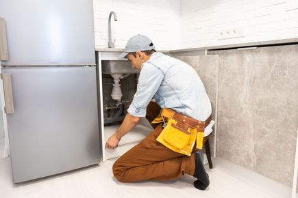 Plumber kneeling under kitchen sink, fixing pipes beside a silver refrigerator and gray tiled wall