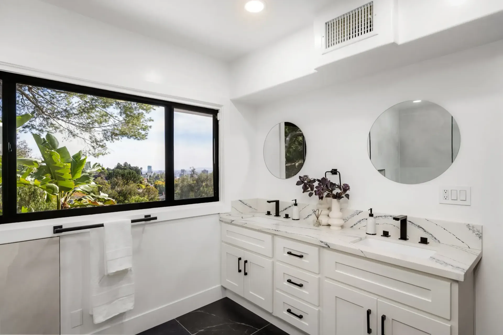 Bright white bathroom with double vanity, round mirrors, and large window overlooking greenery