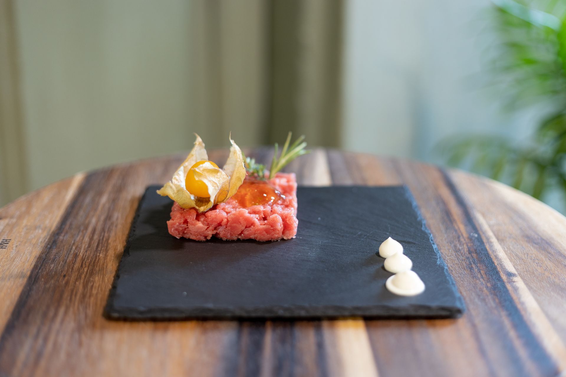 Una fetta quadrata di tartare di manzo rosso guarnita con un frutto di physalis, servita su un piatto di ardesia nera con puntini di panna.