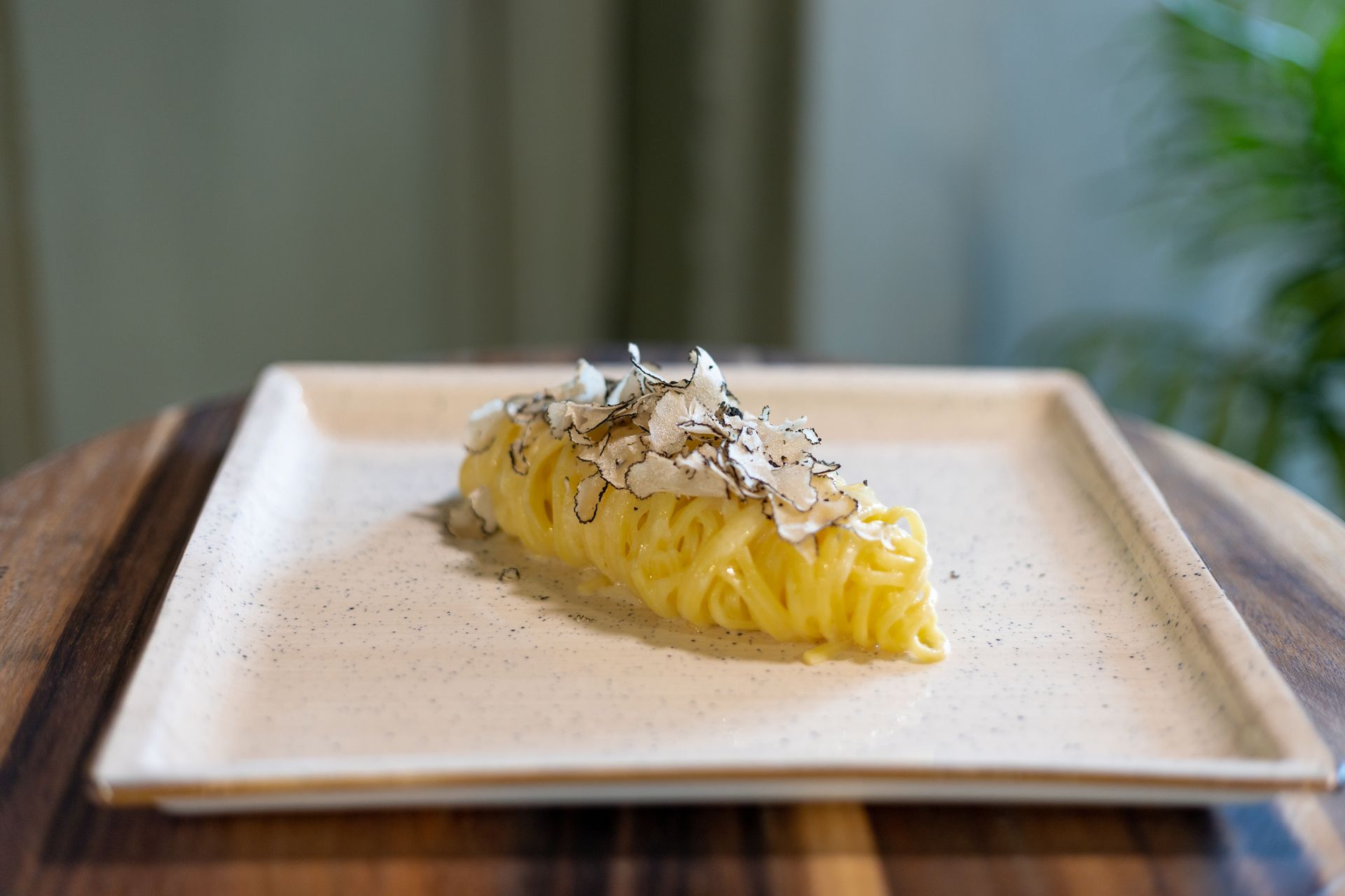 Una porzione di pasta lunga guarnita con scaglie di tartufo, servita su un piatto quadrato di colore chiaro appoggiato su un tagliere di legno.
