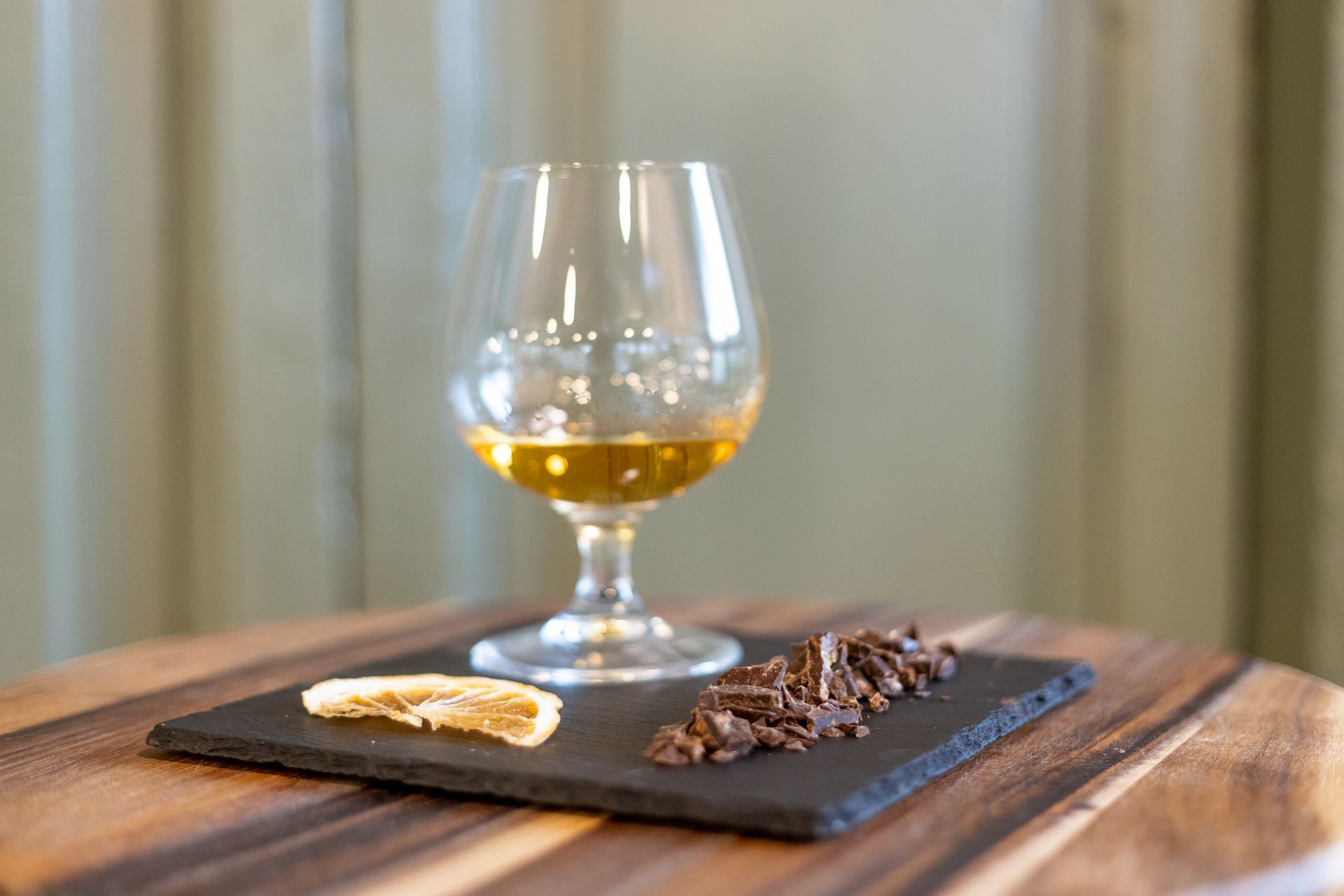 Un bicchiere di liquore color ambra dorata è appoggiato su un tagliere di ardesia accanto a una fetta d'arancia essiccata e a un piccolo mucchietto di gocce di cioccolato fondente.