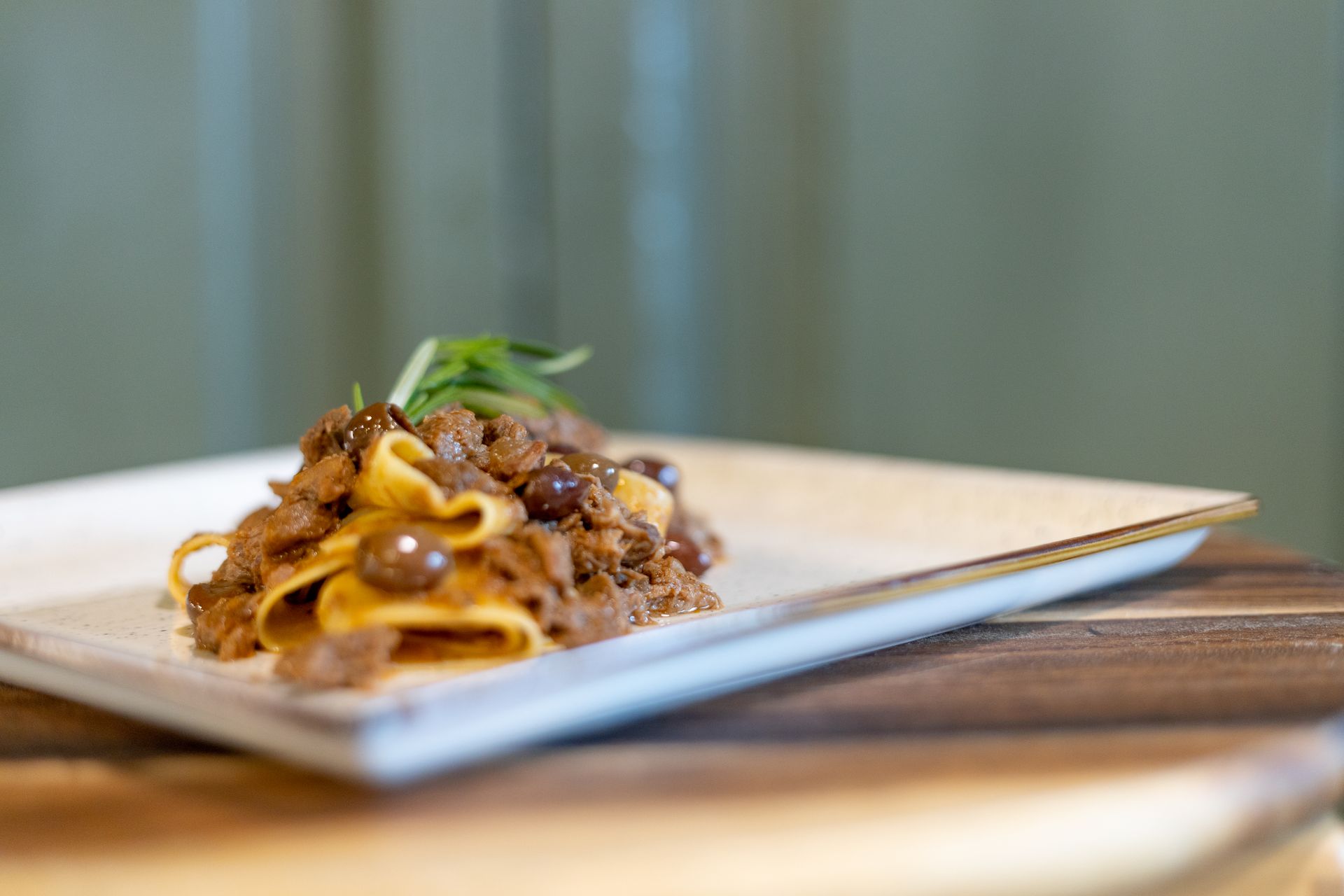 Un piatto di pappardelle con un ricco ragù di carne scura e olive nere, guarnito con erbe aromatiche fresche.