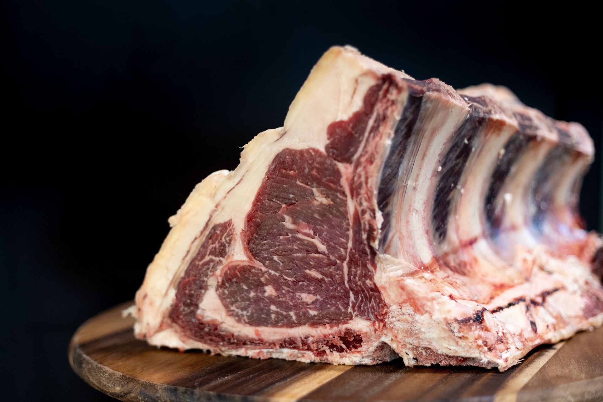 Una costata di manzo cruda con venature di grasso ben visibili, adagiata su un tagliere di legno su uno sfondo scuro.