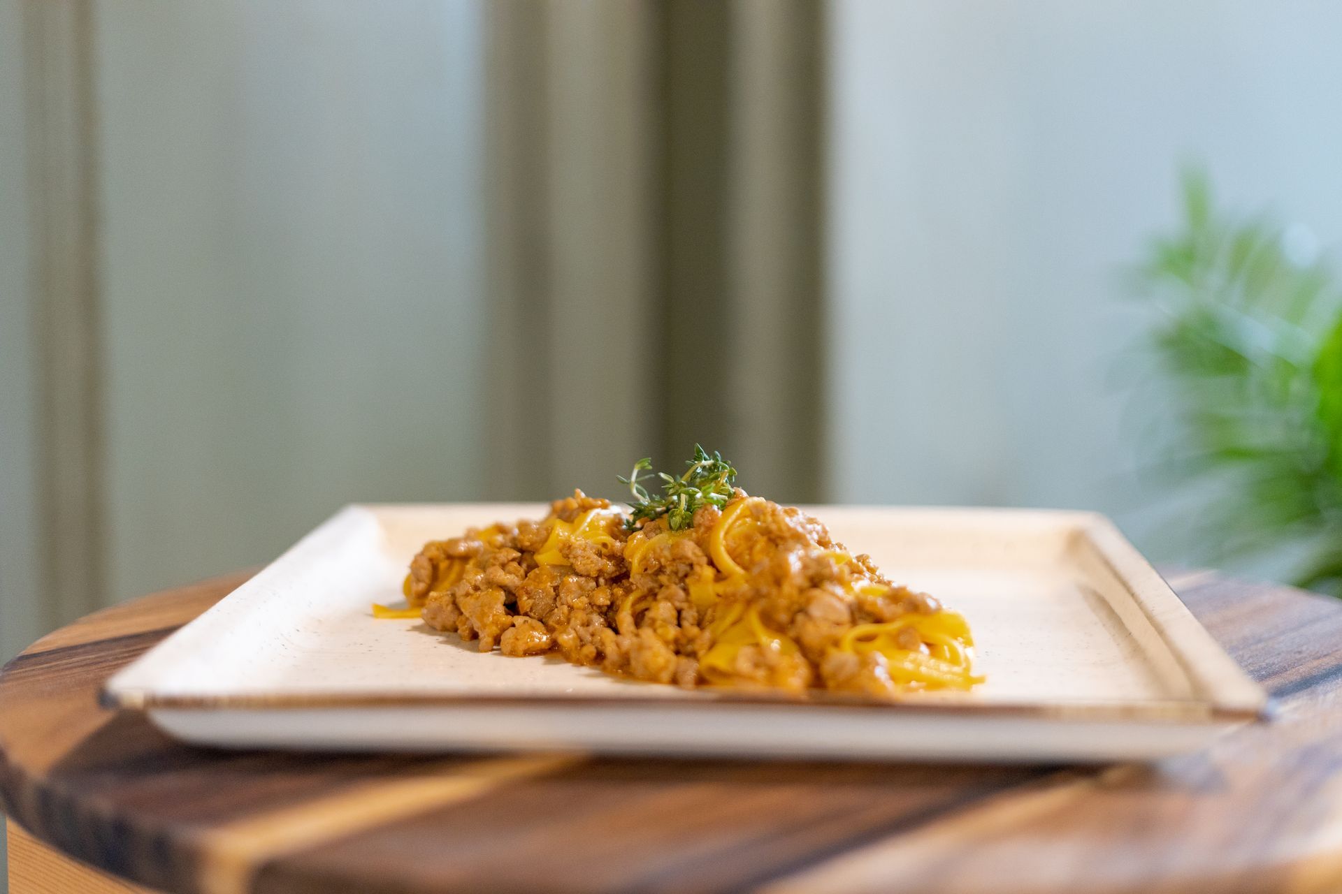 Un piatto di pasta dorata e al sugo, condita con carne macinata e guarnita con erbe aromatiche fresche, adagiato su un vassoio di legno.