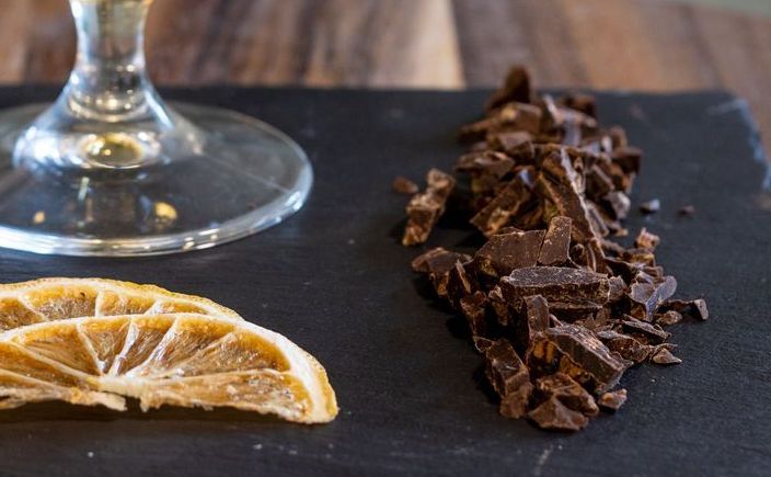 Due fette di agrumi e un mucchietto di cioccolato fondente tritato sono appoggiati su una superficie di ardesia scura accanto a una base di vetro.