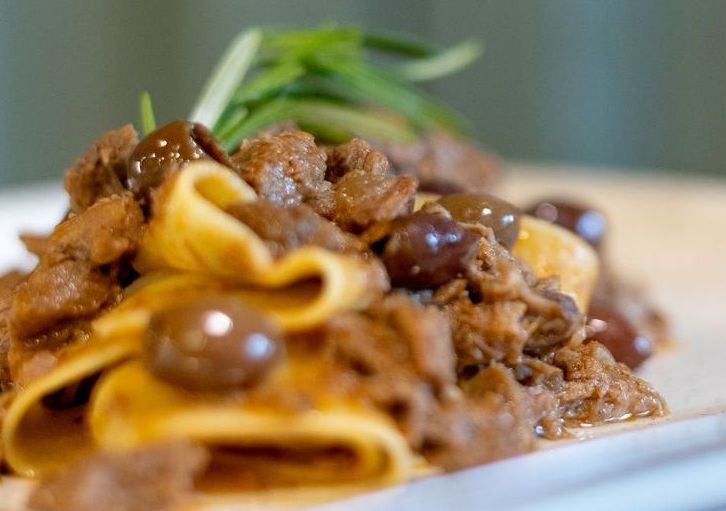Primo piano di pasta con un ricco ragù di carne, guarnita con olive nere e un rametto di erbe aromatiche fresche su un piatto bianco.