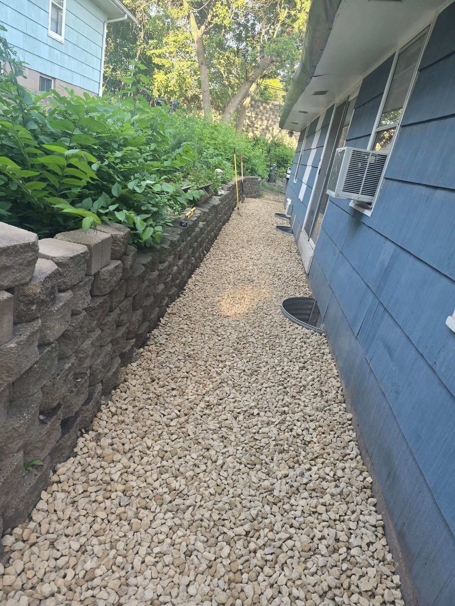 A narrow gravel pathway alongside a blue house and retaining wall with lush green foliage.