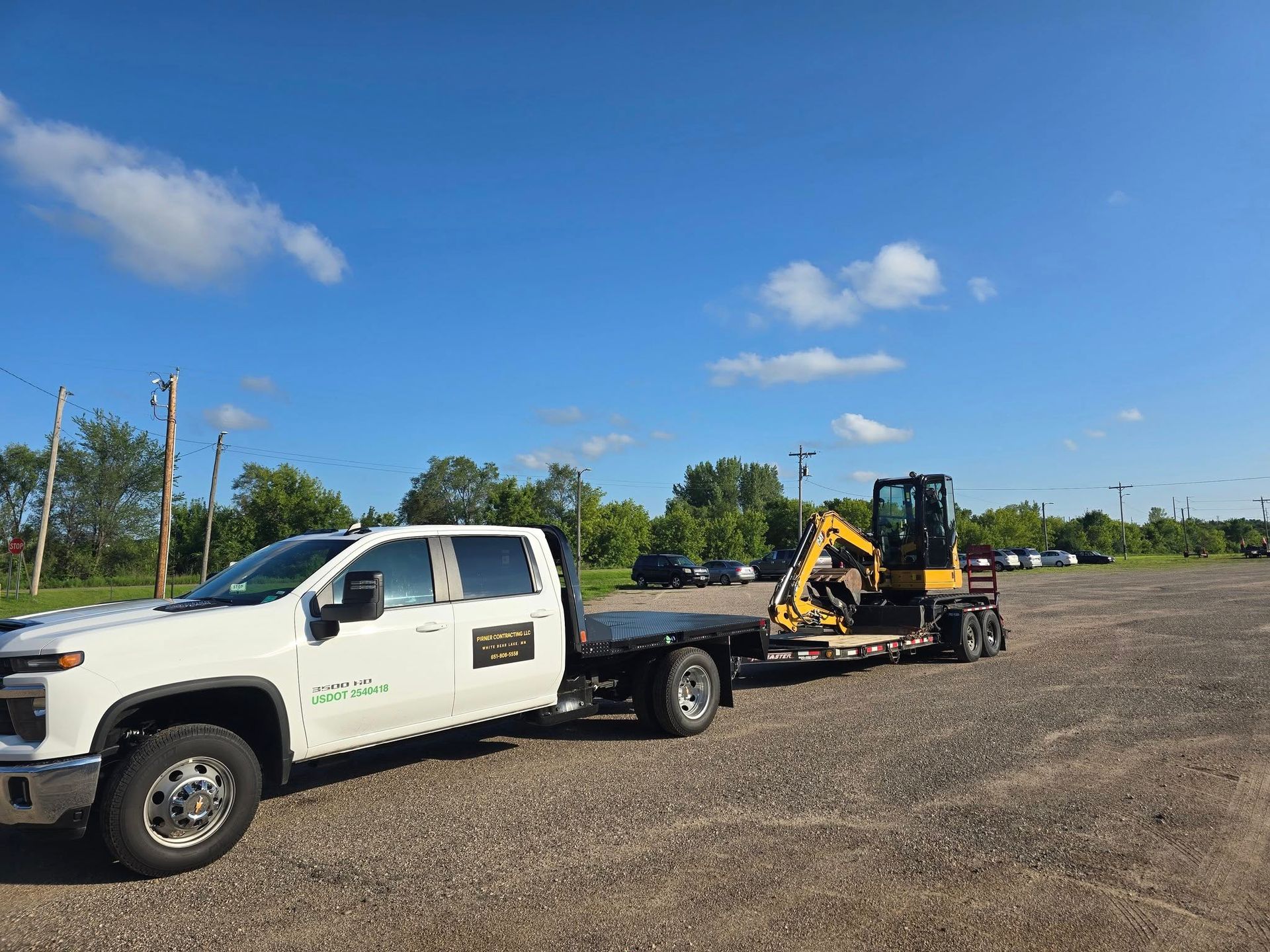 White truck towing a trailer with a yellow excavator on a gravel lot under a blue sky.