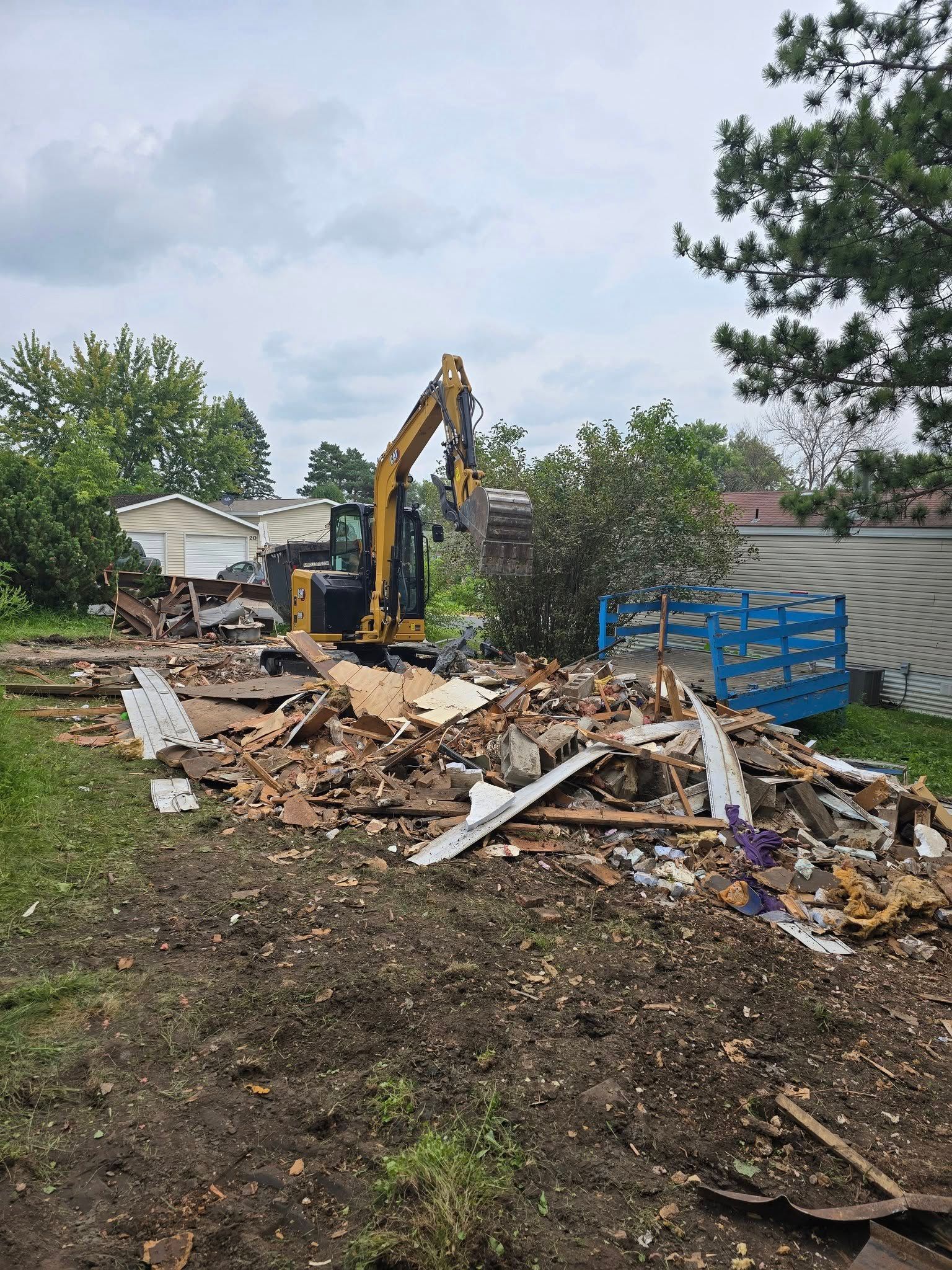 Excavator demolishing a building, surrounded by debris. Overcast sky, green trees, and grass.