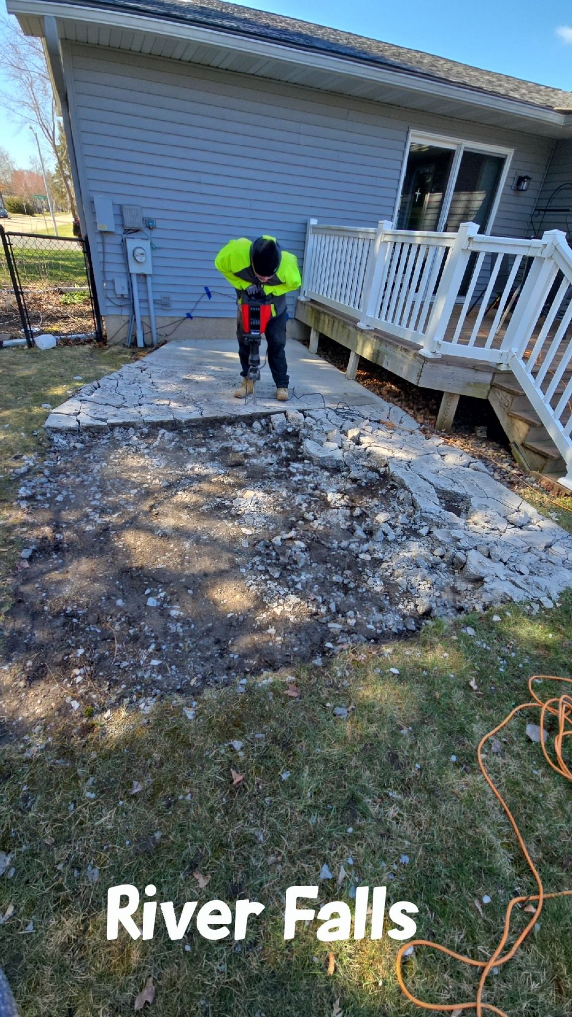 A worker in neon vest uses a jackhammer to remove a concrete patio next to a wooden deck in River Falls.