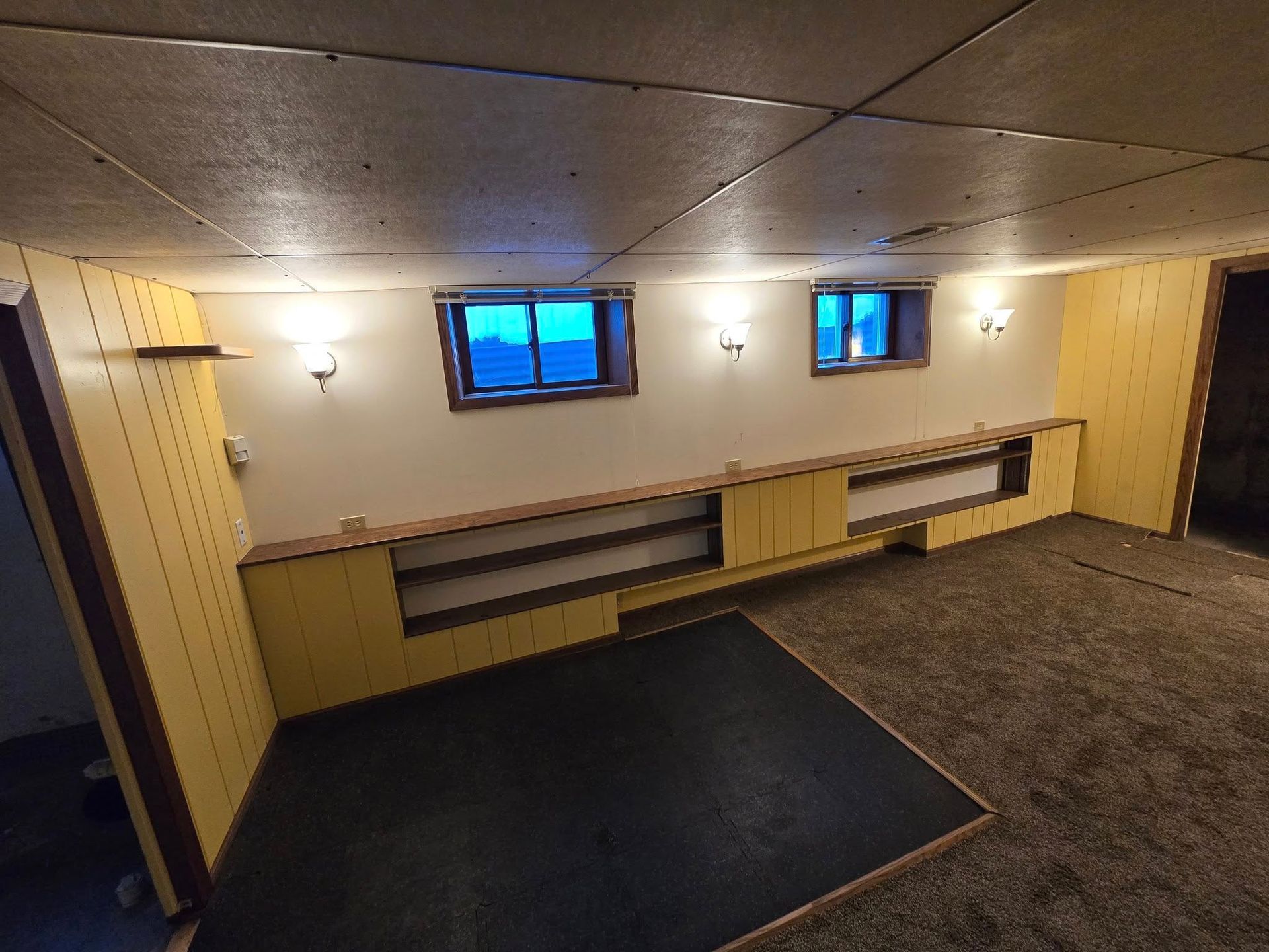Basement interior with two small windows, built-in shelving, and a dark floor area. Yellow walls, tan carpet.