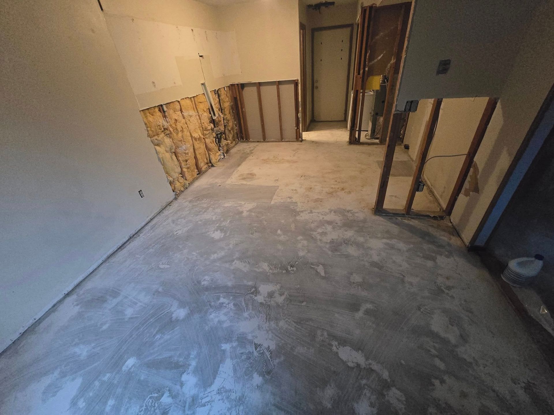 Interior room undergoing construction. Damaged walls with exposed wood framing, concrete floor. Doorway to hallway.