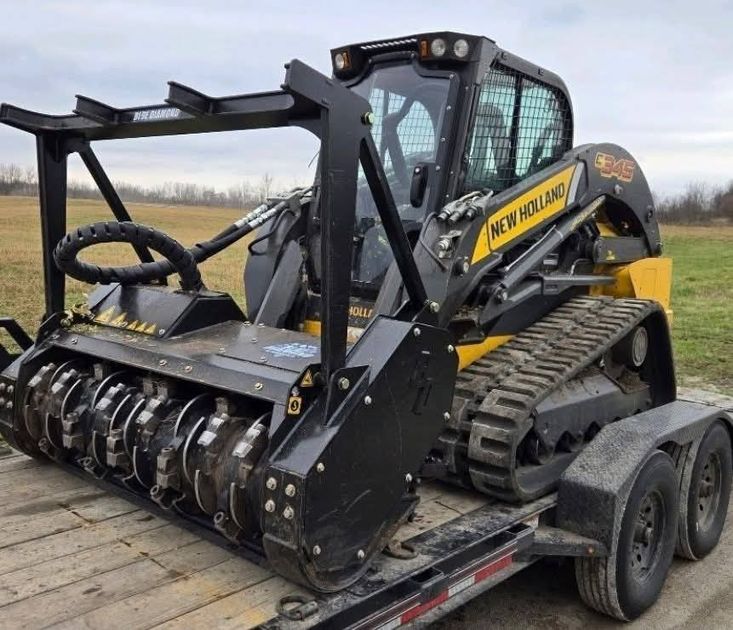 New Holland skid steer with forestry mulcher attachment, on a trailer. Yellow and black machine in a field.
