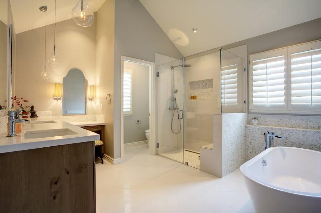 Bright bathroom with double vanity, glass shower, and white soaking tub by shuttered window.