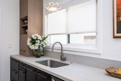 Modern kitchen sink with white countertop, dark cabinets, window, and a vase of white flowers
