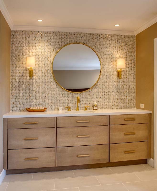 Modern bathroom vanity with wood drawers, round mirror, gold sconces, and mosaic tile backsplash.