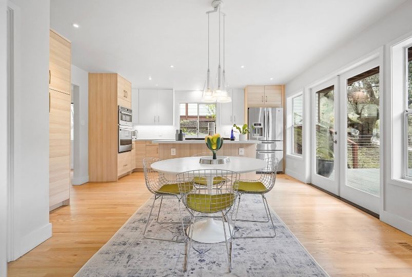 Bright modern dining area with glass table, clear chairs, kitchen, and large windows overlooking greenery