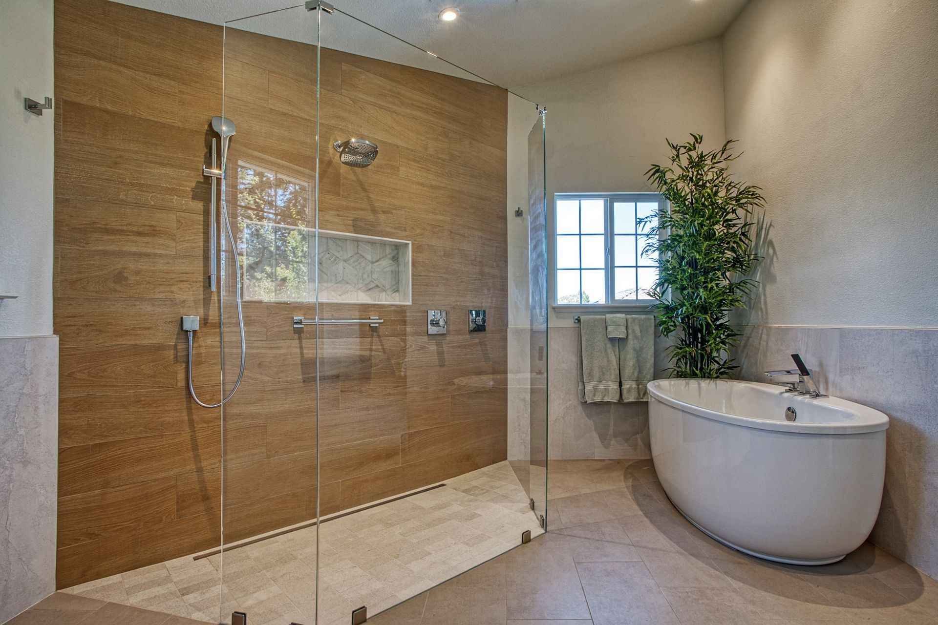 Modern bathroom with glass shower, beige tile, and a freestanding tub by a window.