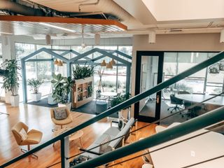Bright modern office lounge with plants, glass meeting pods, and wooden seating overlooking a workspace