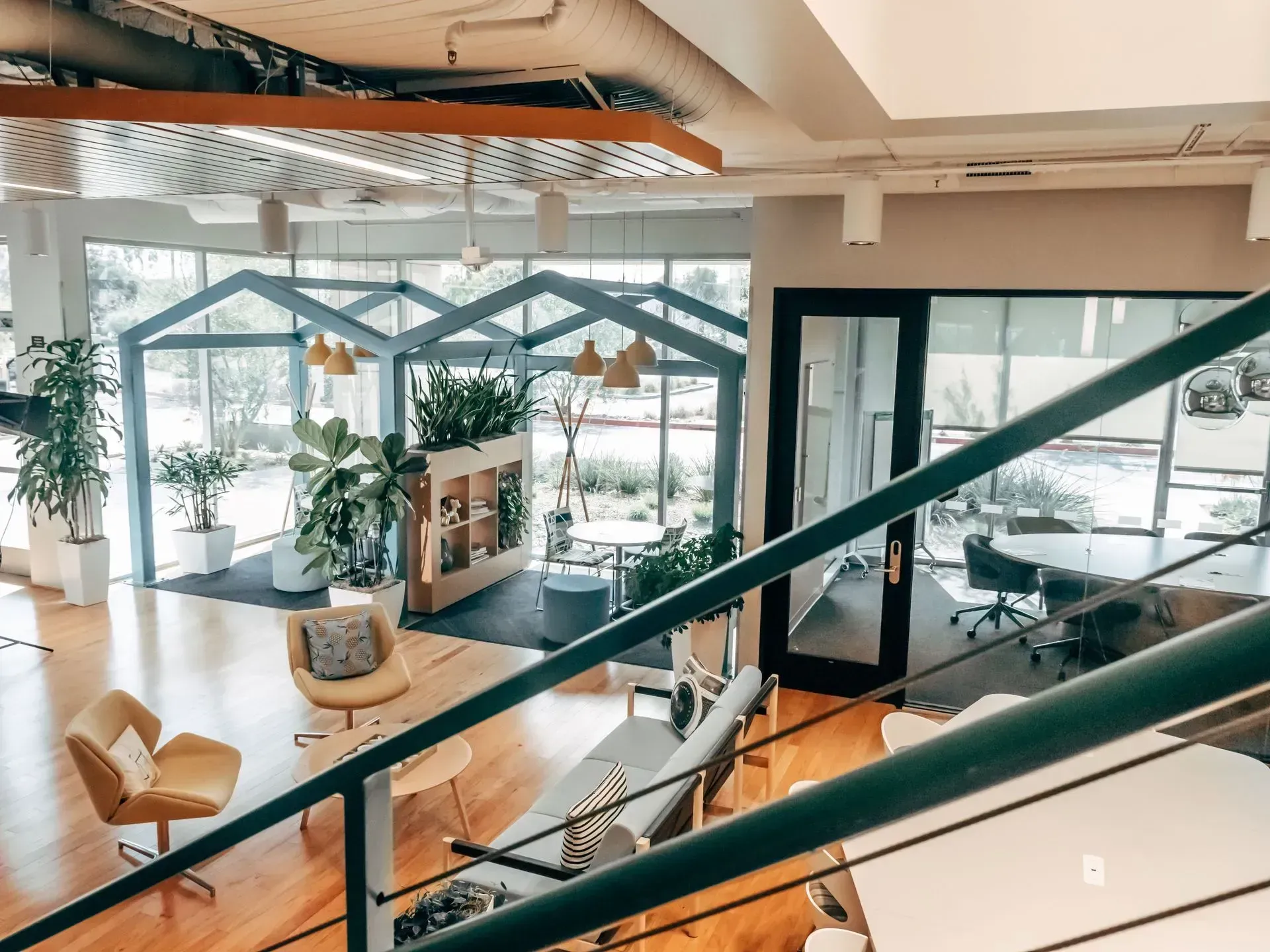 Bright modern office lounge with plants, glass meeting pods, and wooden seating overlooking a workspace