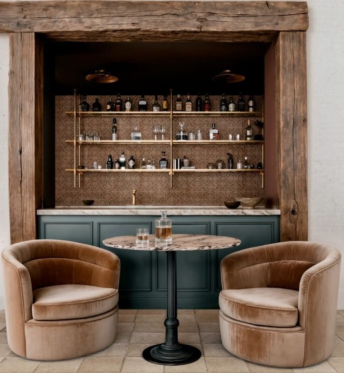 Cozy bar nook with two tan velvet chairs, round table, and shelves of bottles in a rustic wood frame
