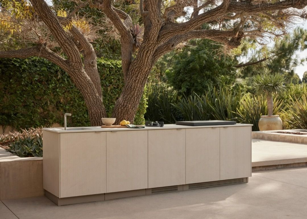 Outdoor kitchen island beneath a large tree in a landscaped garden, with beige cabinetry and potted decor.
