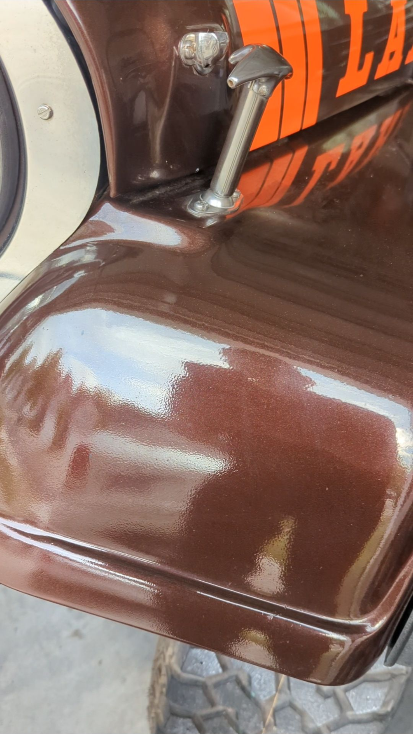 Brown leather motorcycle saddle with a chrome hand lever beside an orange-and-black fuel tank