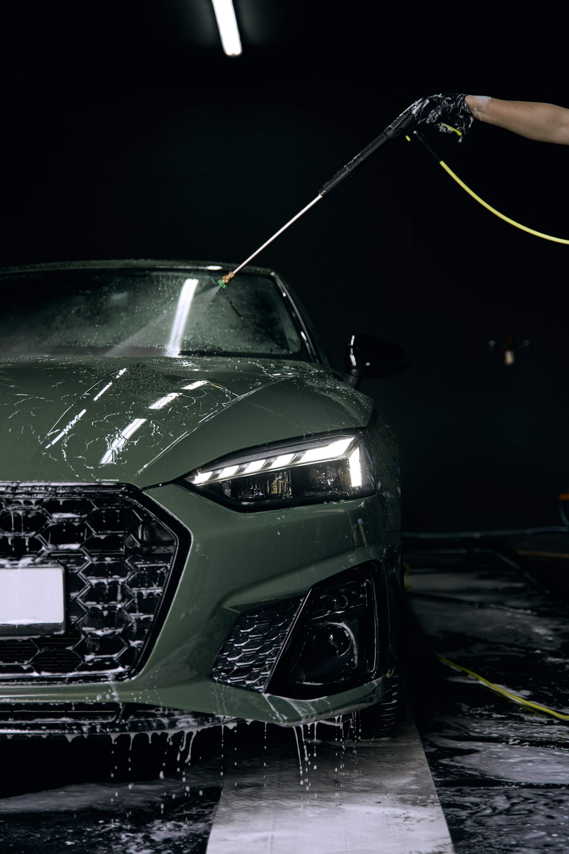 A person washing a dark green car with a water hose in a dimly lit car wash.