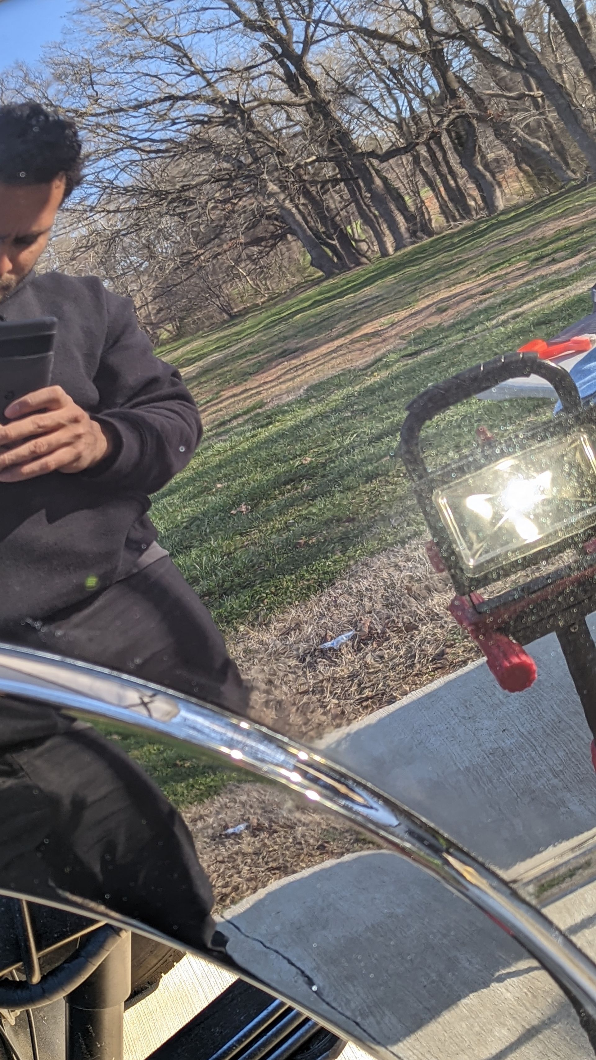 Person in dark clothing taking a mirror selfie outdoors beside a red scooter on a sunny path
