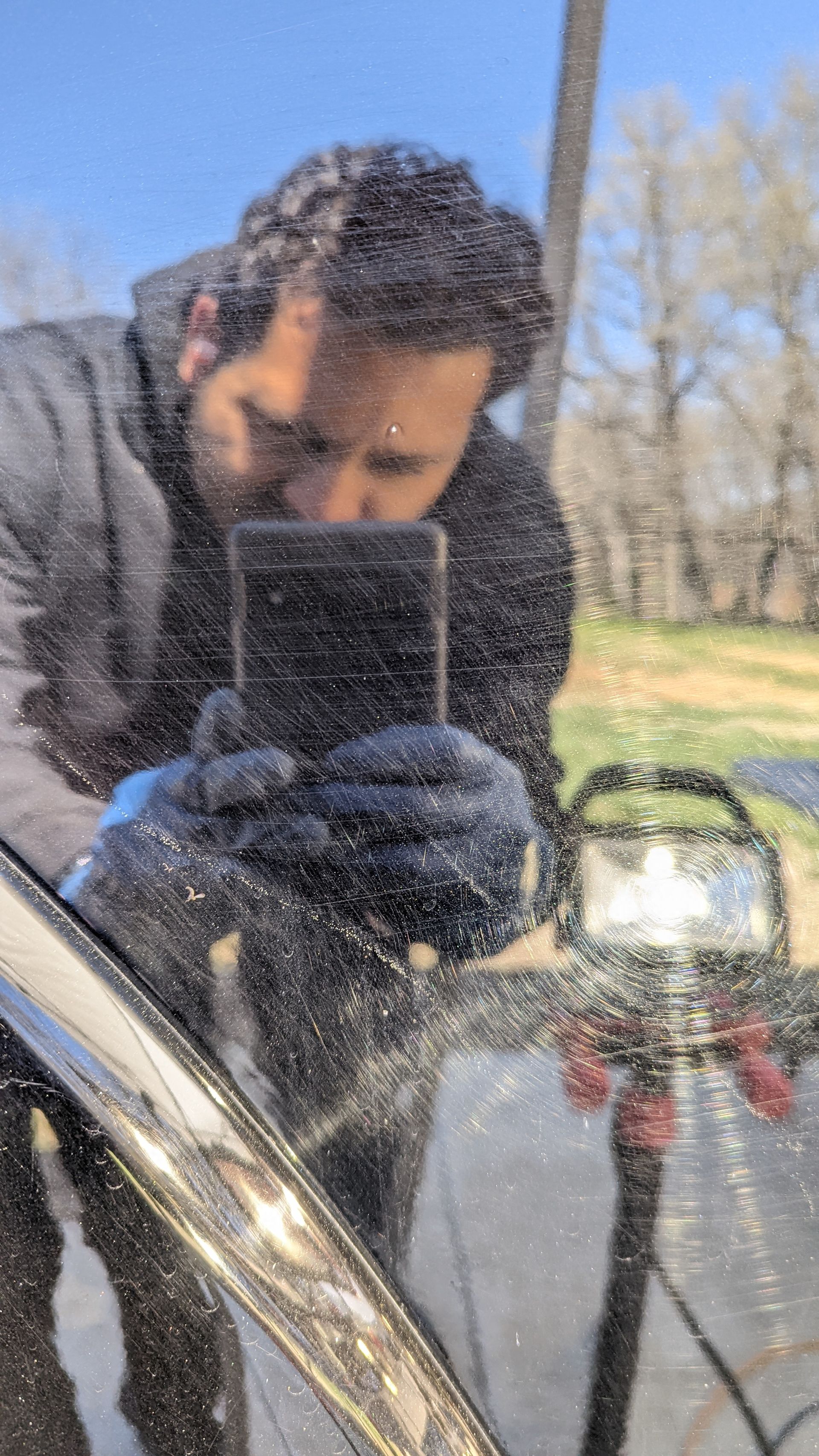 Person in a dark jacket and gloves taking a selfie in a car mirror outdoors on a sunny day