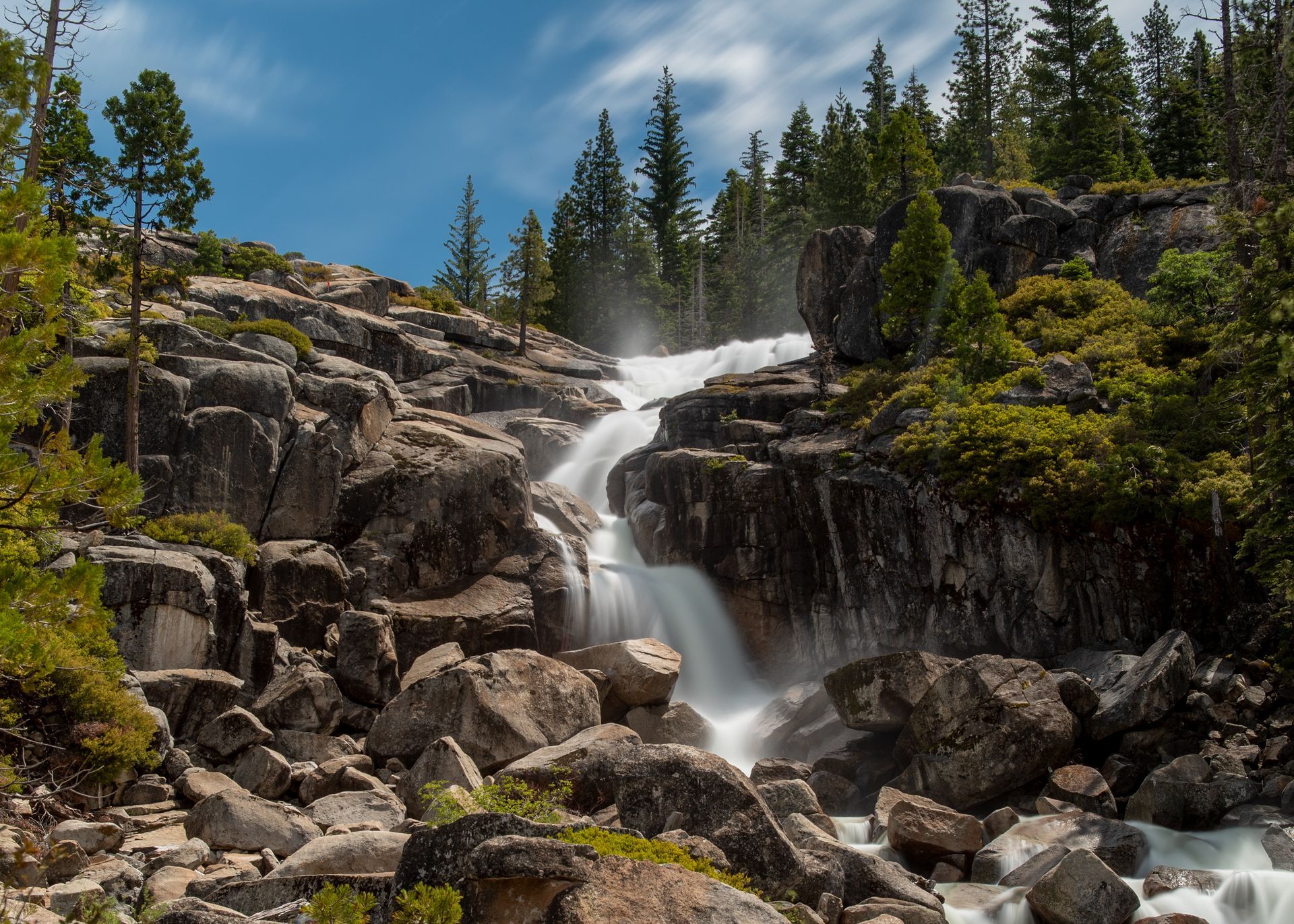 Mountain waterfall cascading over rocky cliffs beneath a blue sky, surrounded by evergreen trees and mossy boulders