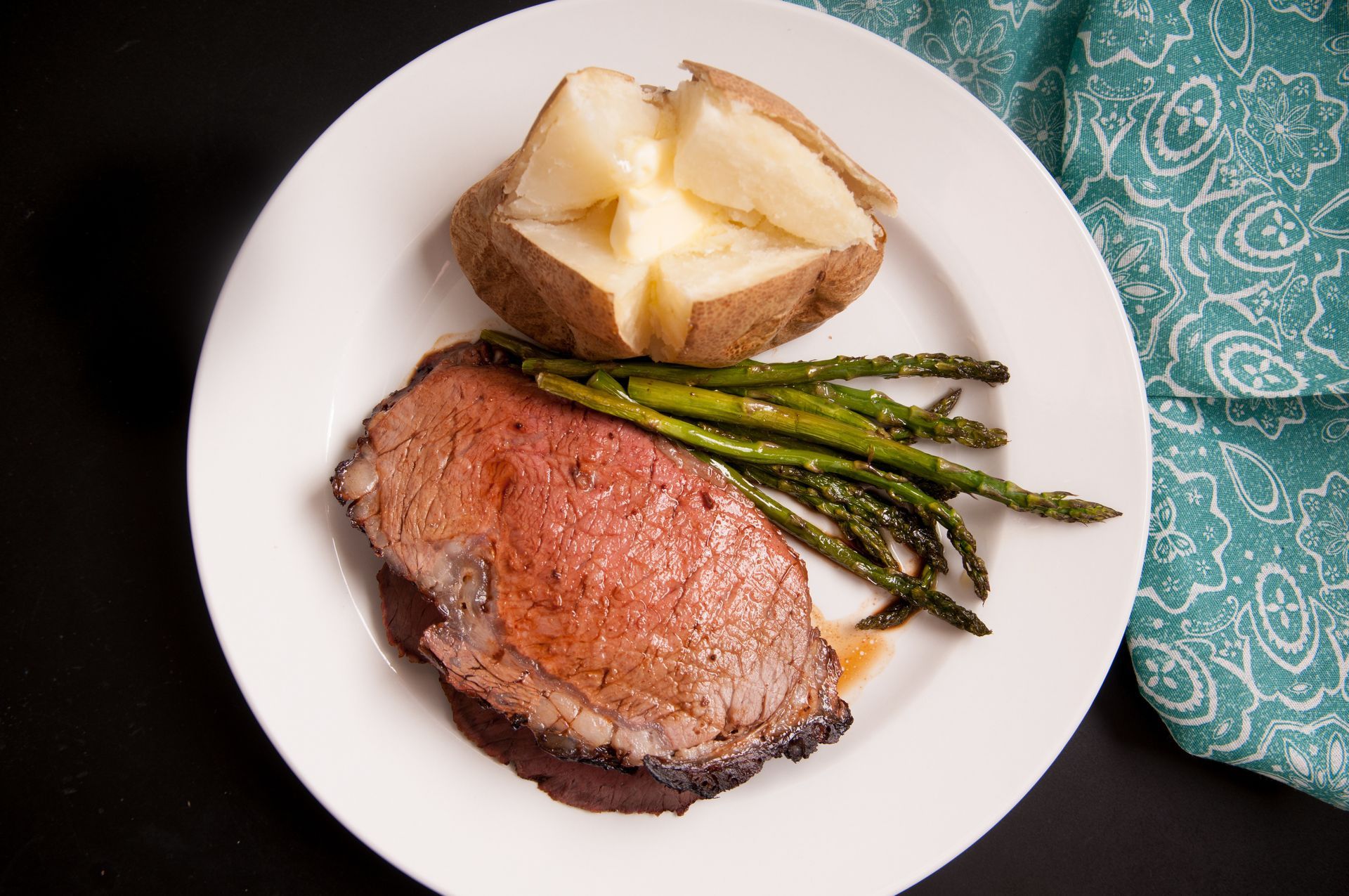 Steak dinner with baked potato and asparagus on a white plate on a dark table