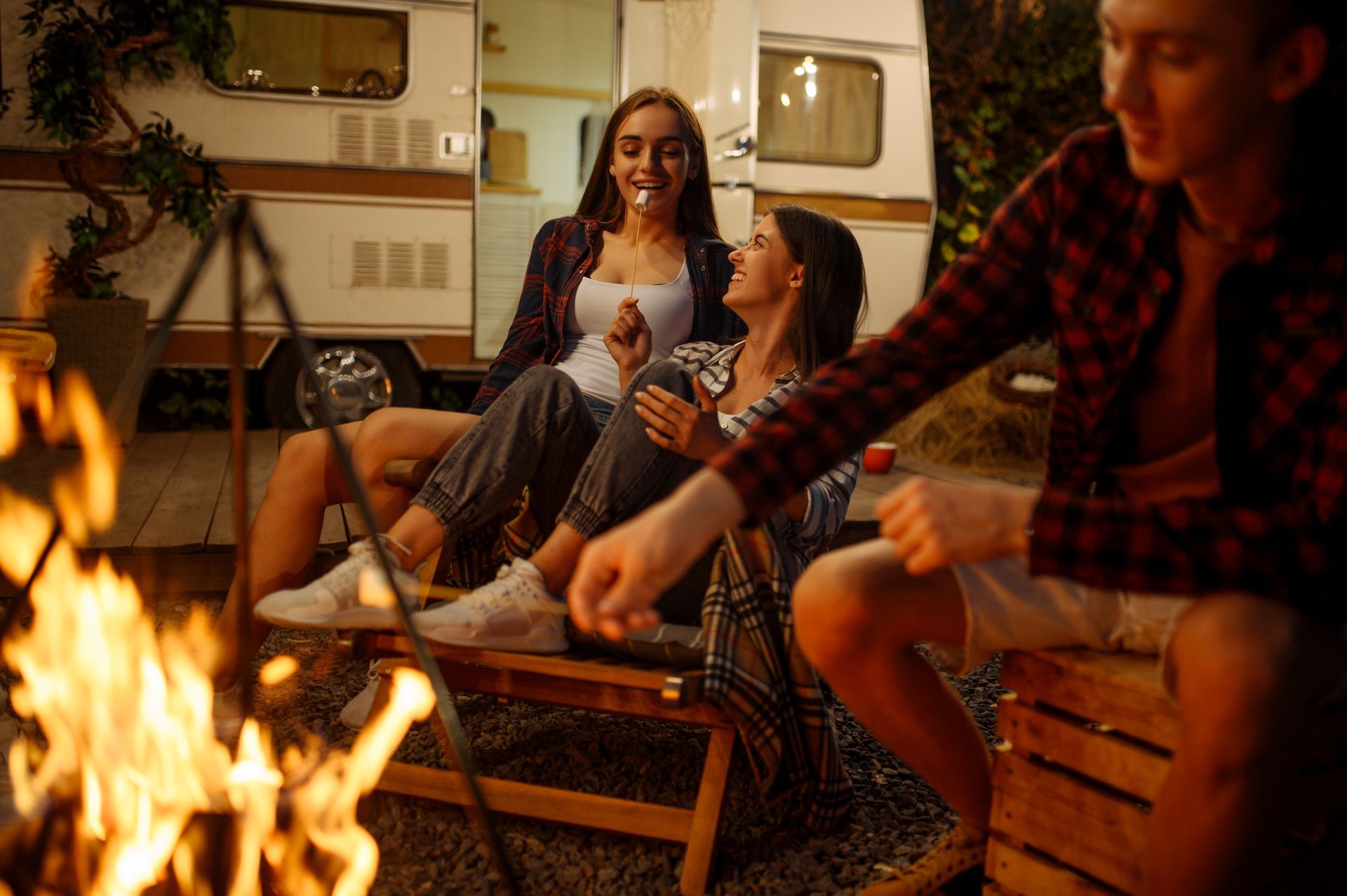 Friends relaxing by a campfire outside a camper, laughing and roasting marshmallows at dusk