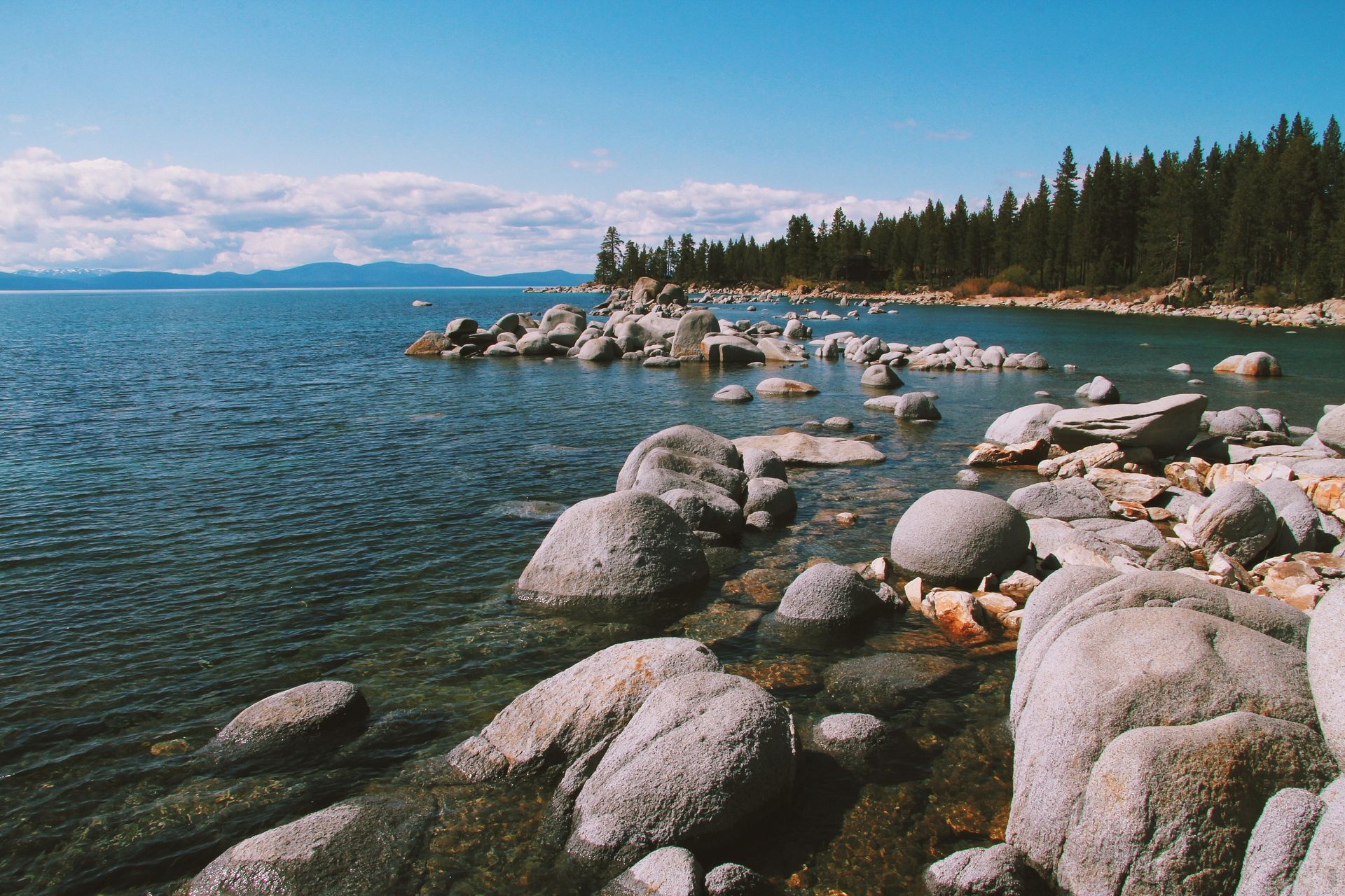 Rocky lakeshore with clear blue water, boulders, pine trees, and distant mountains under a bright sky