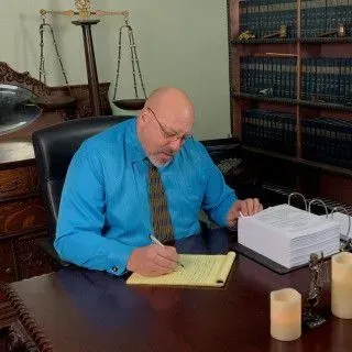 A man in a blue shirt and tie is sitting at a desk writing on a piece of paper.