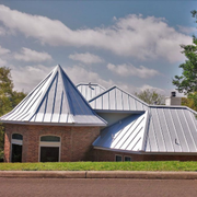 A brick house with a distinctive metal roof, featuring a tall turret and multiple slopes, set near trees under a blue sky.