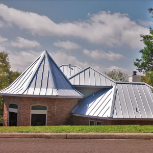 A brick house with a distinctive metal roof, featuring a tall turret and multiple slopes, set near trees under a blue sky.