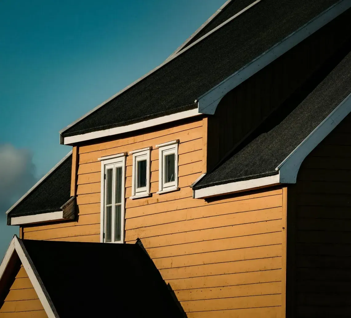 A tan-colored wooden house with black roof shingles stands under a blue sky with soft, partial clouds.