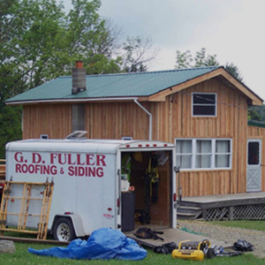 A GD Fuller Roofing & Siding trailer parked next to a wood-sided cabin with a green metal roof.