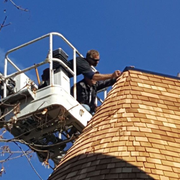 Workers in a lift bucket installing wood shingles on the curved roof of a Victorian-style house under a blue sky.
