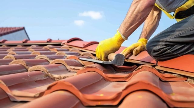 A worker wearing yellow gloves uses a tool to install or repair terracotta roof tiles on a sunny day.