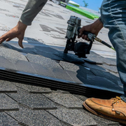 A worker in work clothes and yellow gloves uses a tool to install dark asphalt shingles on a residential roof.