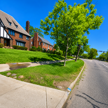 Sunny suburban street with brick houses, green lawn, and a tree beside a curving road