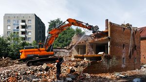 Orange excavator demolishing a brick house amid rubble, with an apartment building in the background