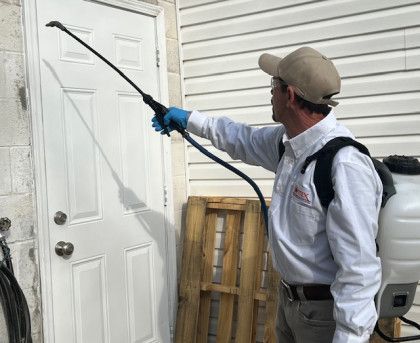 Pest control worker spraying a white door. He wears a hat, gloves, and a backpack sprayer.