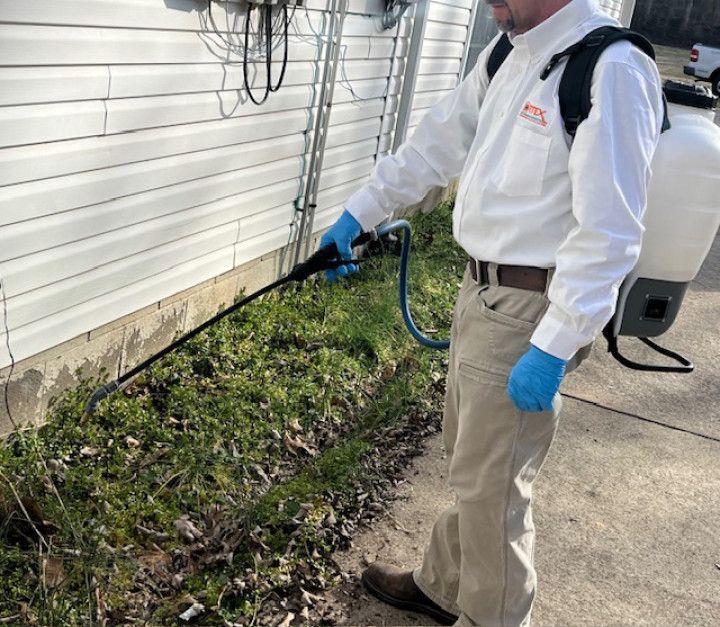 Pest control worker spraying herbicide near a building. White uniform, gloves, backpack sprayer.
