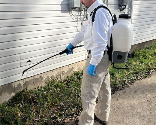 Pest control worker spraying insecticide along a house foundation with a backpack sprayer