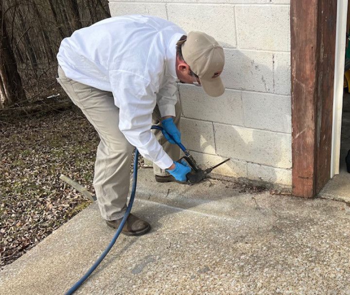 Man in light-colored clothes sprays a building's base with a hose, wearing gloves and a cap.