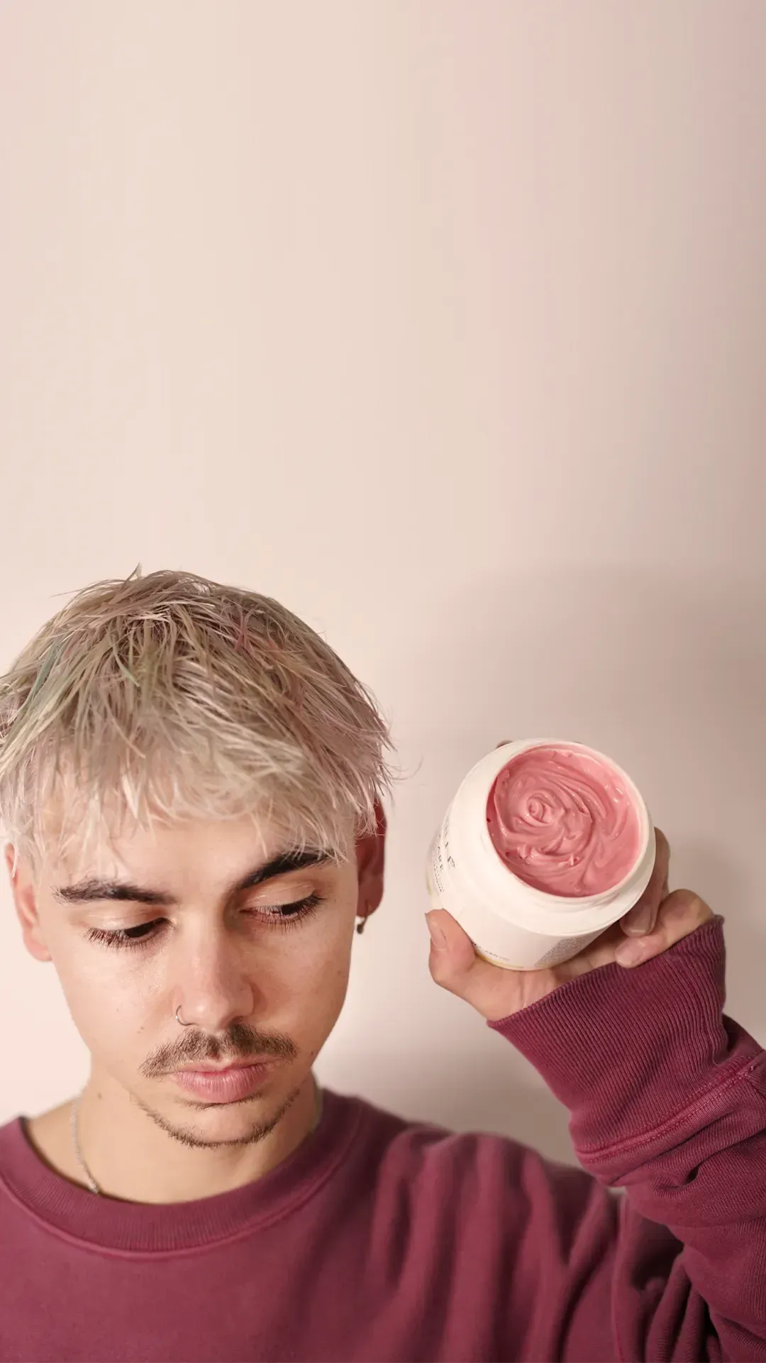 Person with short blond hair holds a pink makeup compact near their face against a pale background.
